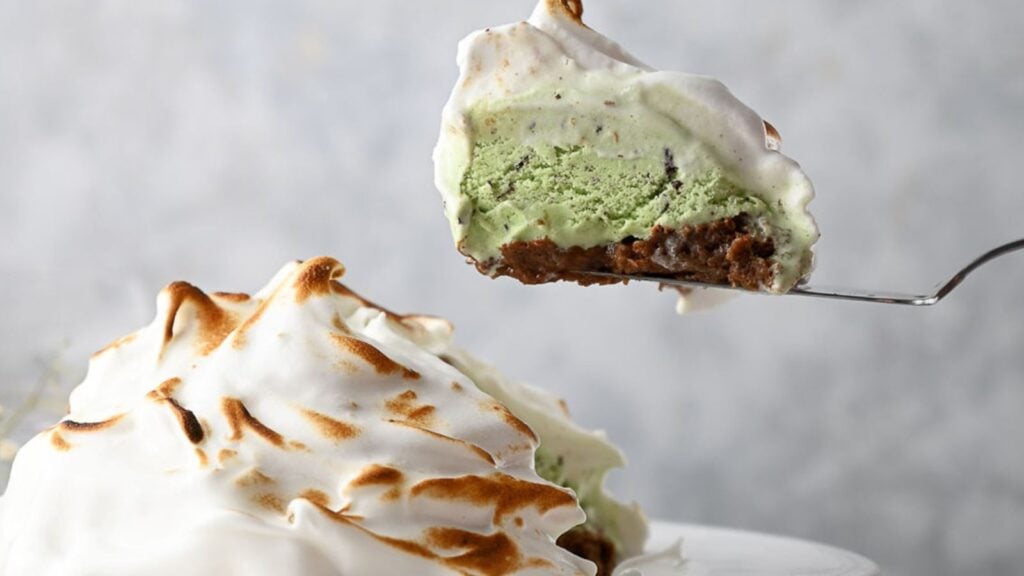 A slice of baked Alaska with a layer of green ice cream, cake base, and toasted meringue topping on a utensil, with the remainder in the background.