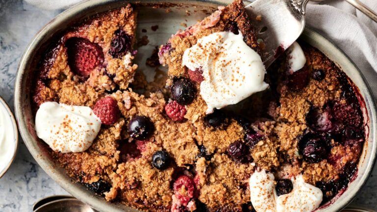 Baked berry oatmeal in an oval dish, topped with dollops of cream and sprinkled cinnamon, with a serving spoon lifting a portion.