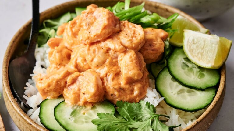 A bowl of rice topped with shrimp in a creamy sauce, garnished with cucumber slices, lime wedge, cilantro, and sliced green onions.