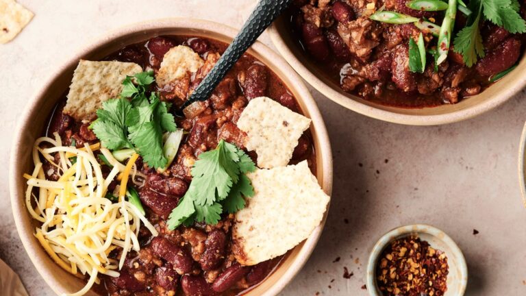 Bowls of chili with kidney beans, shredded cheese, tortilla chips, and cilantro garnished on top. A small bowl of red chili flakes is on the side.