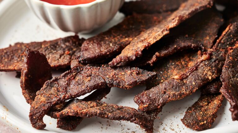 A plate of dark, seasoned beef jerky strips with a small bowl of red dipping sauce beside them.
