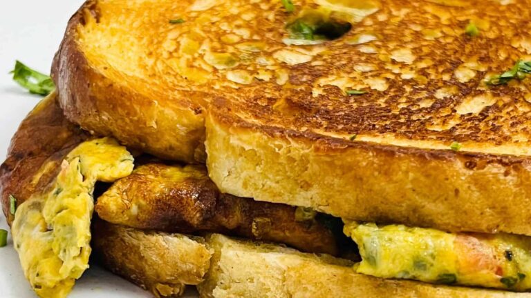 Close-up of a toasted sandwich with scrambled eggs between thick slices of golden brown bread. Garnished with small green herbs.