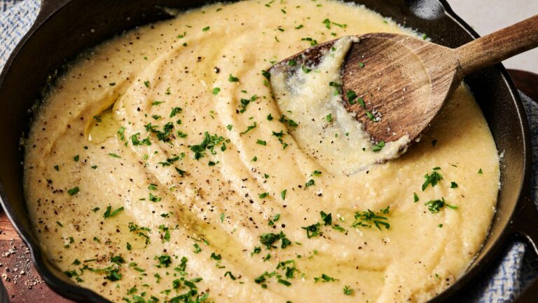 Creamy polenta in a skillet, topped with chopped herbs and black pepper, and stirred with a wooden spoon.
