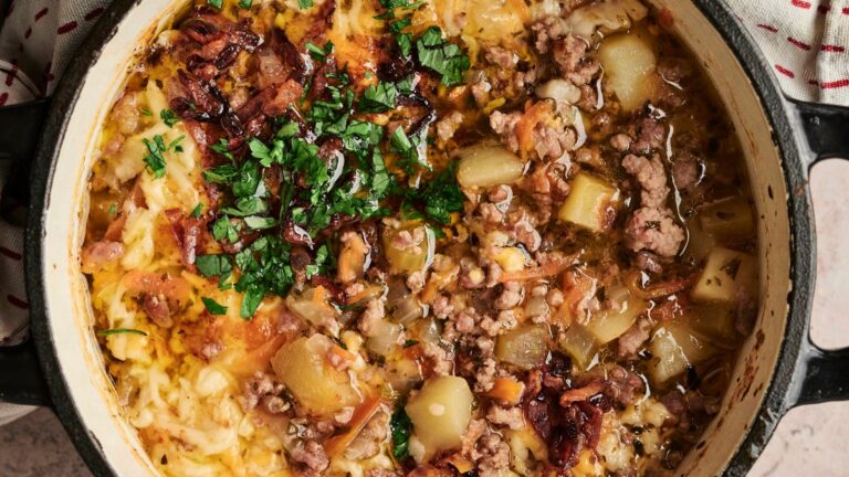 A pot containing a stew of minced meat, diced potatoes, cheese, onions, and greens. Garnished with chopped parsley and crispy bacon bits.