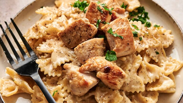 A plate of bowtie pasta with creamy sauce, topped with grilled chicken pieces and garnished with parsley. A black fork rests on the plate.