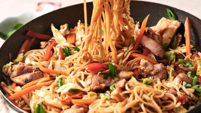 Stir-fried noodles with sliced chicken, carrots, onions, and cabbage in a pan.
