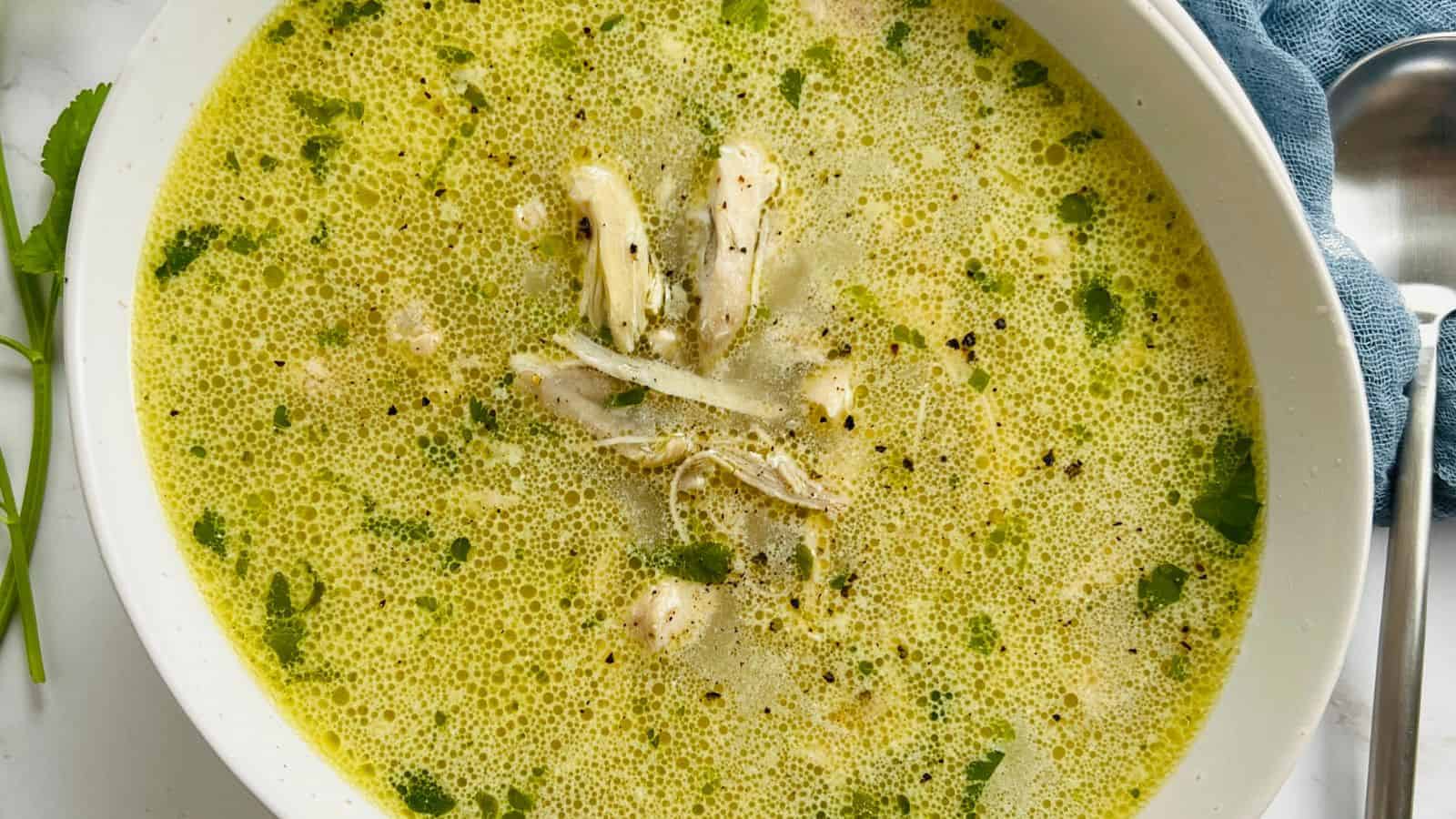 A bowl of chicken soup with herbs and black pepper, next to a blue cloth and a spoon.