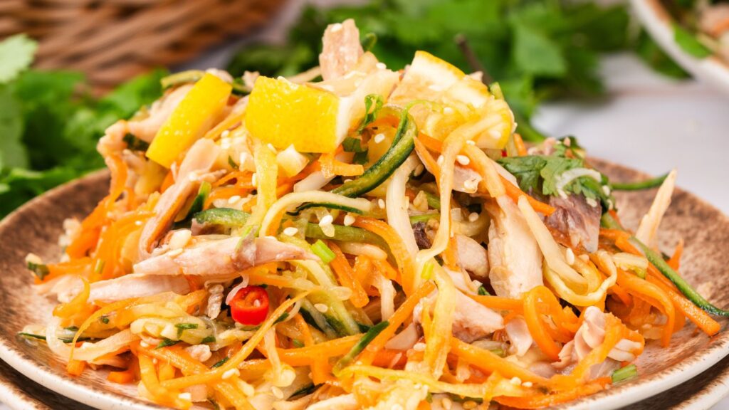 A plate of Asian-style salad with shredded vegetables, chicken, orange slices, sesame seeds, and garnished with herbs.