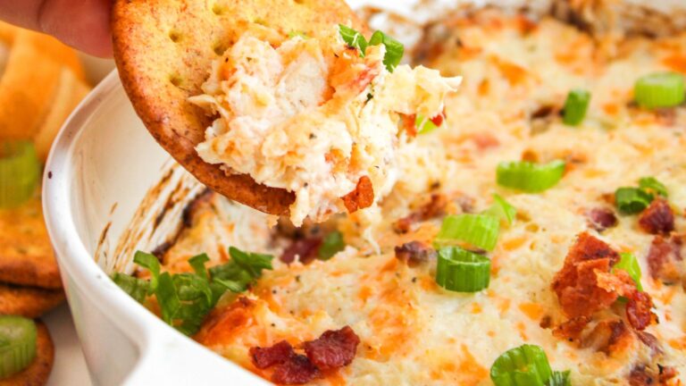 Cracker with creamy dip featuring bacon bits and green onions being lifted from a dish.