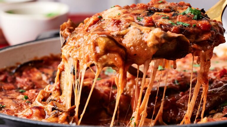 A serving of cheesy eggplant parmesan being lifted from a pan, with melted cheese stretching and herbs sprinkled on top.