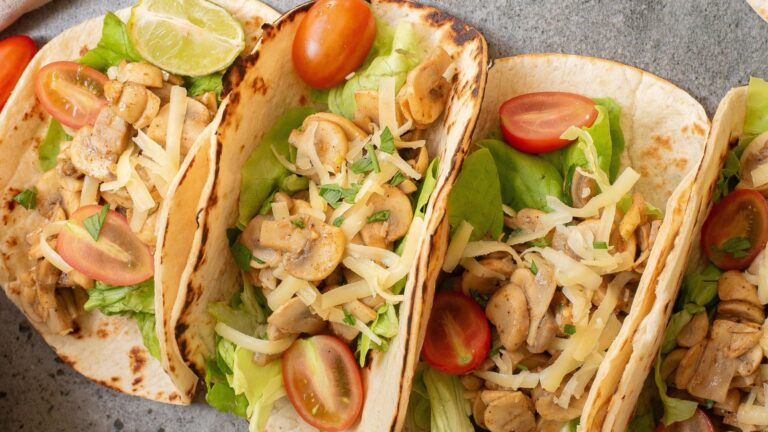 Three tacos filled with lettuce, sliced mushrooms, grated cheese, and cherry tomato halves. A lime wedge is on the side.