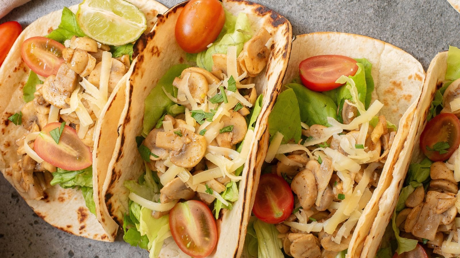 Three soft tacos filled with lettuce, mushrooms, shredded cheese, and cherry tomatoes, garnished with a lime wedge.