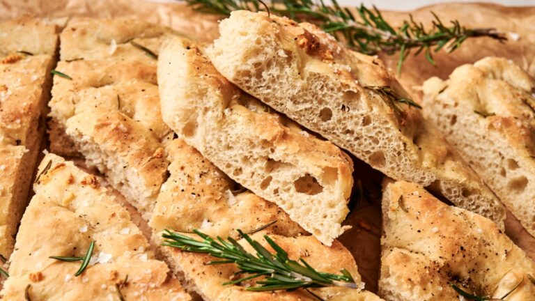 Slices of focaccia bread topped with rosemary and flaky salt, arranged on brown parchment paper.