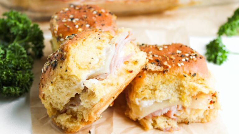 Three ham and cheese sliders on seeded buns are on display, surrounded by fresh parsley.