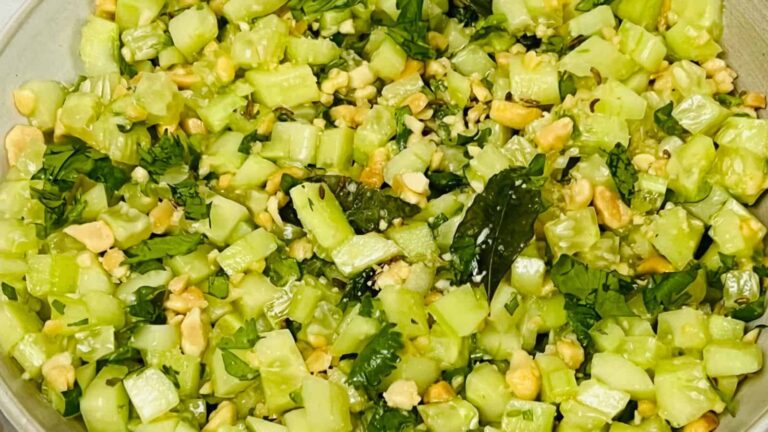 A close-up of a green salad with chopped cucumbers, herbs, and nuts in a bowl.