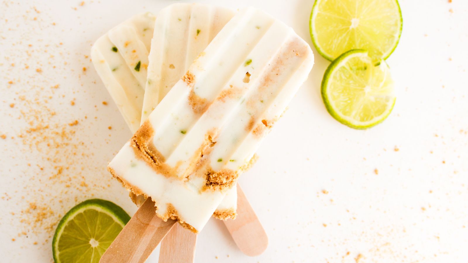Three creamy popsicles with lime slices and crumbs scattered around on a white surface.