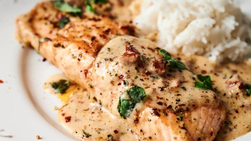 Grilled salmon fillet with creamy sauce and herbs, served alongside a portion of white rice on a plate.