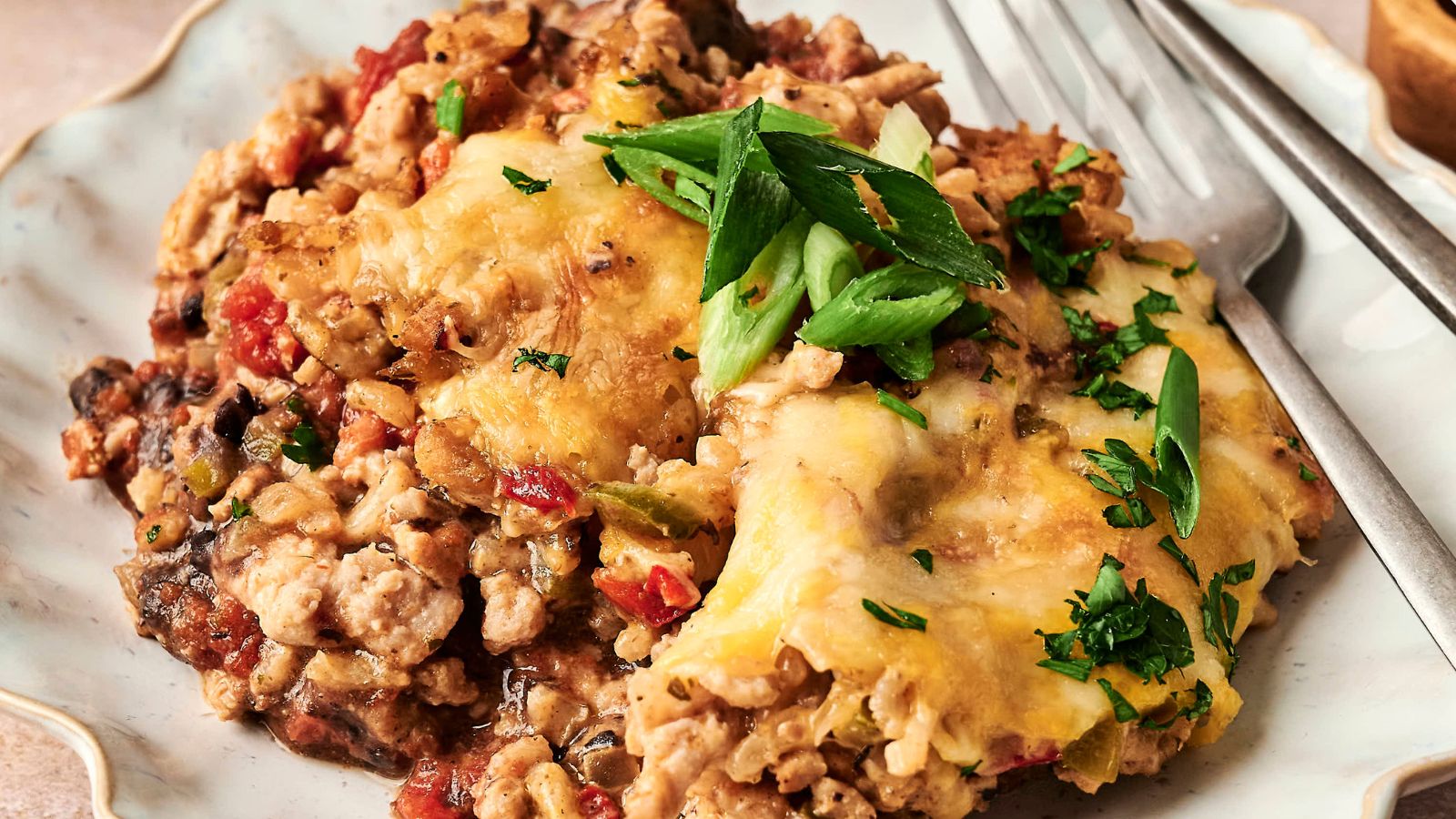 A plated serving of cheesy casserole with ground meat, topped with sliced green onions and parsley, accompanied by a fork.