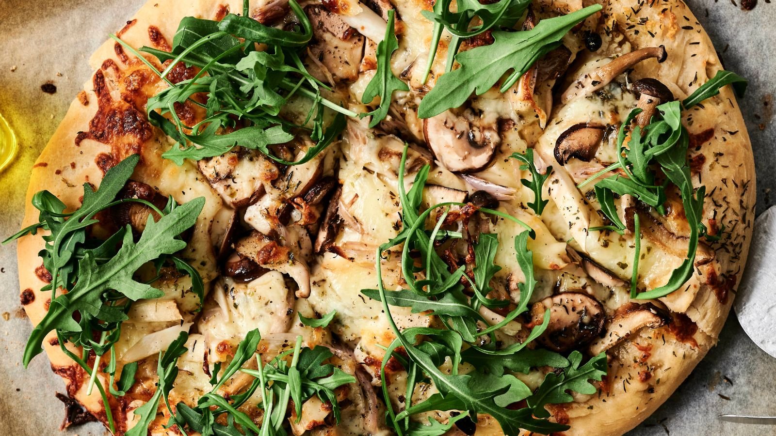 Close-up of a pizza topped with arugula, mushrooms, cheese, and herbs on a parchment paper.
