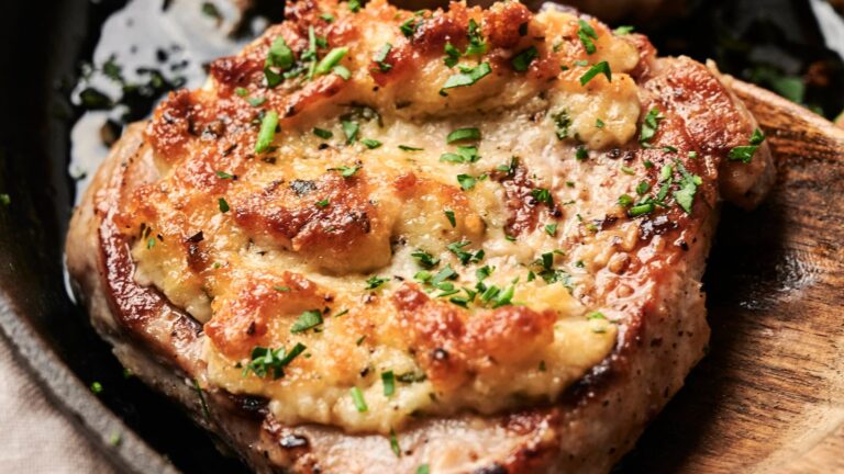 Close-up of a cooked, seasoned pork chop topped with melted cheese and garnished with chopped parsley on a dark plate.