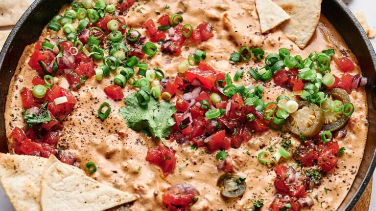A skillet with cheesy dip topped with chopped tomatoes, green onions, jalapeños, and cilantro, surrounded by tortilla chips.