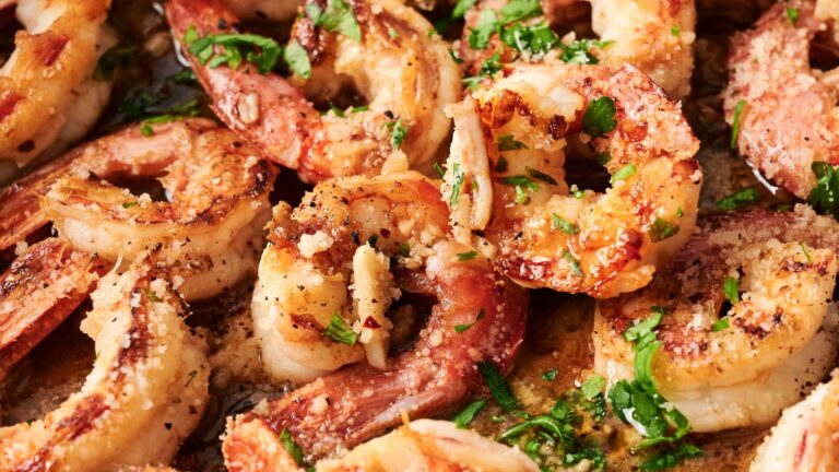 Close-up of grilled shrimp garnished with chopped herbs on a plate. The shrimp are seasoned with visible spices and cooked to a golden brown.