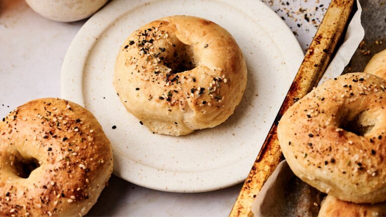 A plate with a sesame-seed bagel next to a baking tray holding several more bagels.