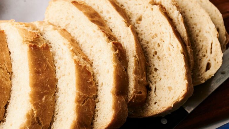 Sliced loaf of beige bread on a wooden cutting board, with a bread knife partially visible.