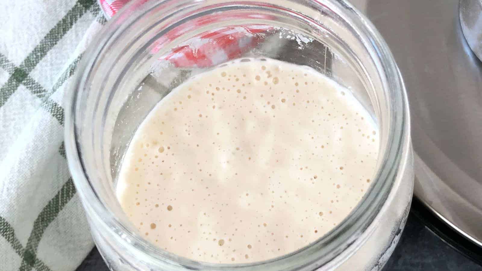 Open glass jar filled with frothy, light-beige liquid next to a green and white checkered cloth.