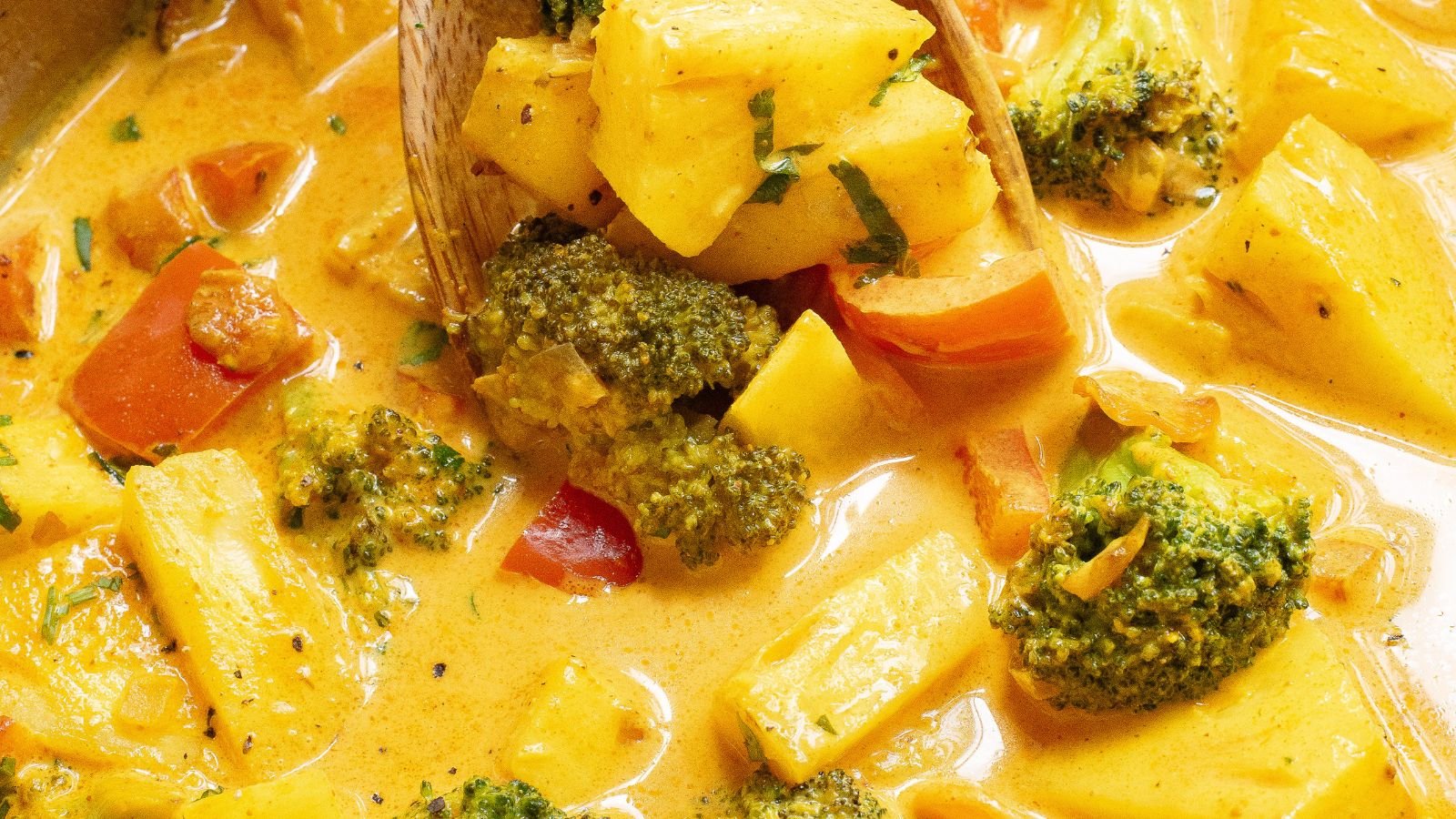 Close-up of a creamy yellow curry dish with broccoli, pineapple, and red bell peppers, stirred with a wooden spoon.