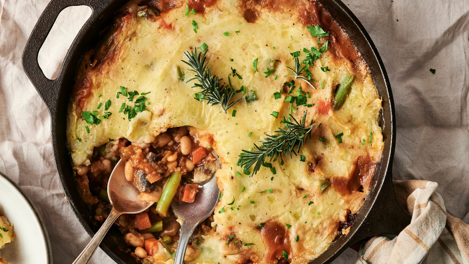 A cast iron skillet filled with vegetable shepherd's pie, topped with mashed potatoes and garnished with rosemary sprigs and chopped parsley. Two spoons are placed inside.