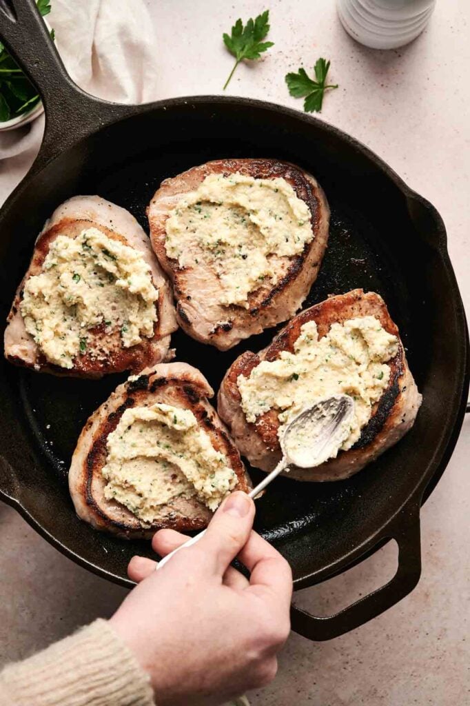 A hand delicately applies a creamy herb spread onto pork chops sizzling in a cast iron skillet, infusing them with flavor.
