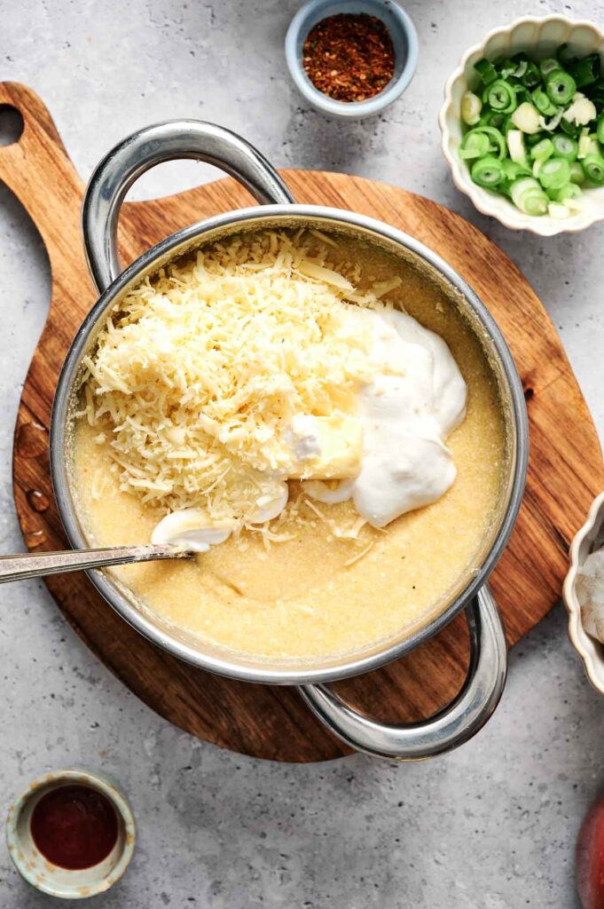 A pot of shrimp and grits with shredded cheese and sour cream sits on a wooden board, surrounded by small bowls of chopped green onions and spices.