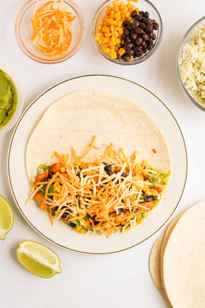 A tortilla on a plate topped with shredded cheese, beans, corn, and vegetables, surrounded by bowls of rice, beans with corn, cheese, and guacamole, plus lime wedges.