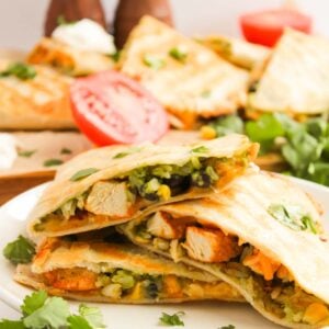 Quesadillas filled with vegetables and chicken are stacked on a white plate, surrounded by cilantro and tomato slices.