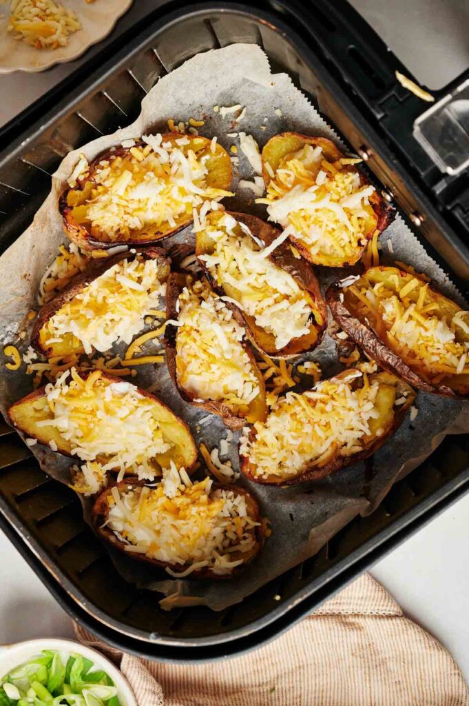 Air Fryer Loaded Potato Skins are topped with shredded cheese and cooked to crispy perfection in an air fryer lined with parchment paper. A dish scattered with chopped green onions adds a fresh touch to the savory delight visible at the bottom.