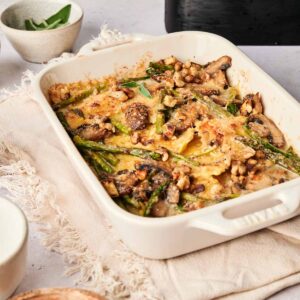This air fryer ravioli casserole features baked pasta with melted cheese, mushrooms, asparagus, and walnuts. Served in a rectangular dish, it's accompanied by a small bowl of walnuts and herbs on a beige cloth for a cozy dining experience.