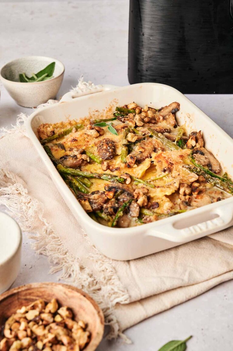 This air fryer ravioli casserole features baked pasta with melted cheese, mushrooms, asparagus, and walnuts. Served in a rectangular dish, it's accompanied by a small bowl of walnuts and herbs on a beige cloth for a cozy dining experience.