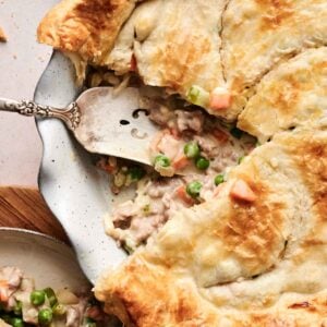 Close-up of an air fryer beef pot pie with a slice removed, revealing a savory filling of peas, carrots, and tender meat. A metal spatula rests in the dish, ready to serve another slice onto the nearby plate.