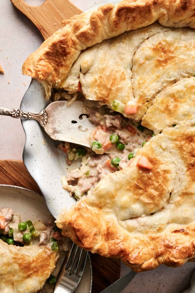 Close-up of an air fryer beef pot pie with a slice removed, revealing a savory filling of peas, carrots, and tender meat. A metal spatula rests in the dish, ready to serve another slice onto the nearby plate.