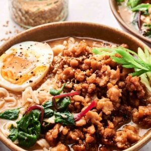 A bowl of chicken ramen with minced meat, half a boiled egg, spinach, and greens. Black chopsticks and a jar with seasoning are in the background.