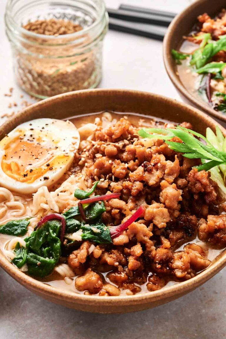 A bowl of chicken ramen with minced meat, half a boiled egg, spinach, and greens. Black chopsticks and a jar with seasoning are in the background.