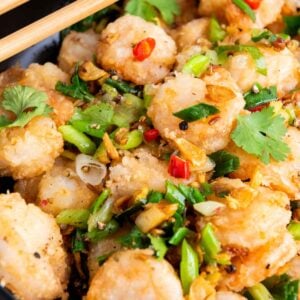 A bowl of fried shrimp garnished with chopped green onions, red chilies, cilantro, and black sesame seeds, with chopsticks resting on the edge.