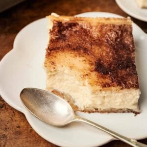 A square piece of churro cheesecake with a cinnamon topping on a white plate, next to a silver spoon.