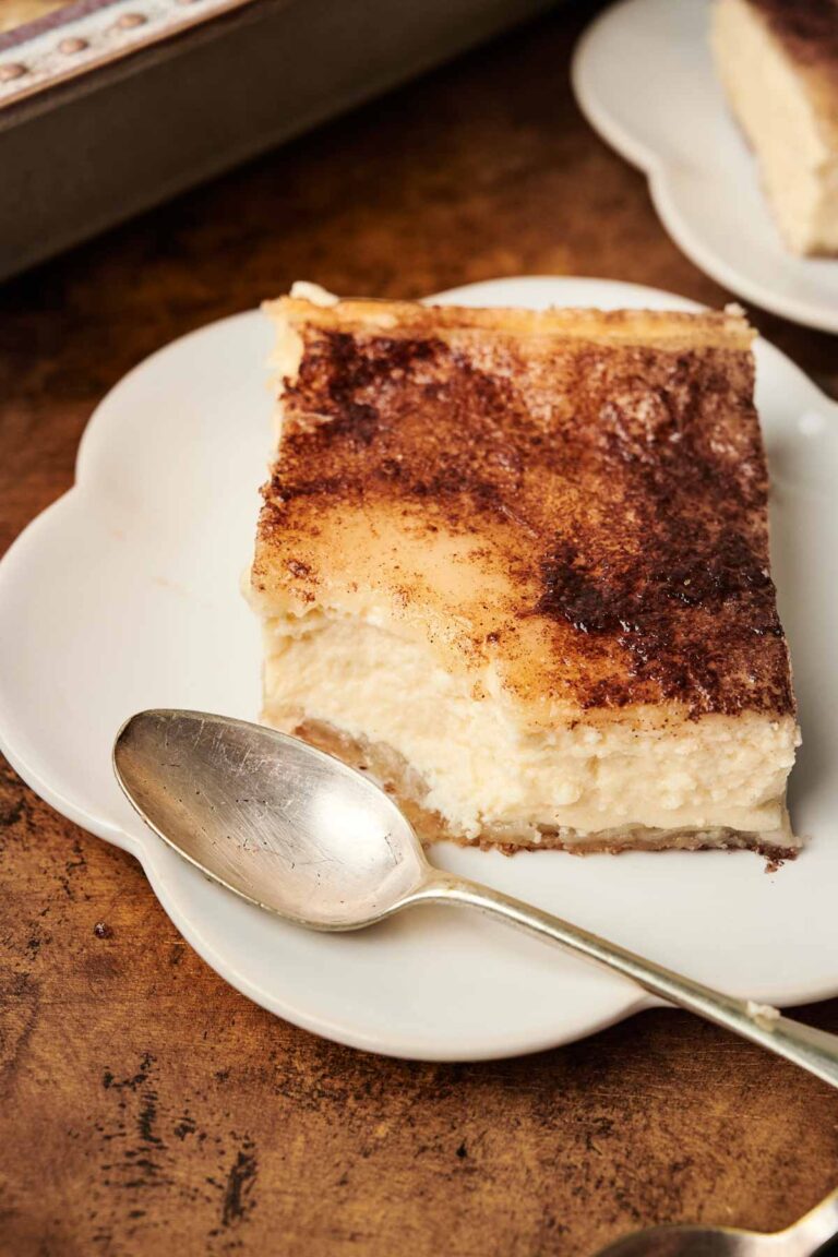 A square piece of churro cheesecake with a cinnamon topping on a white plate, next to a silver spoon.