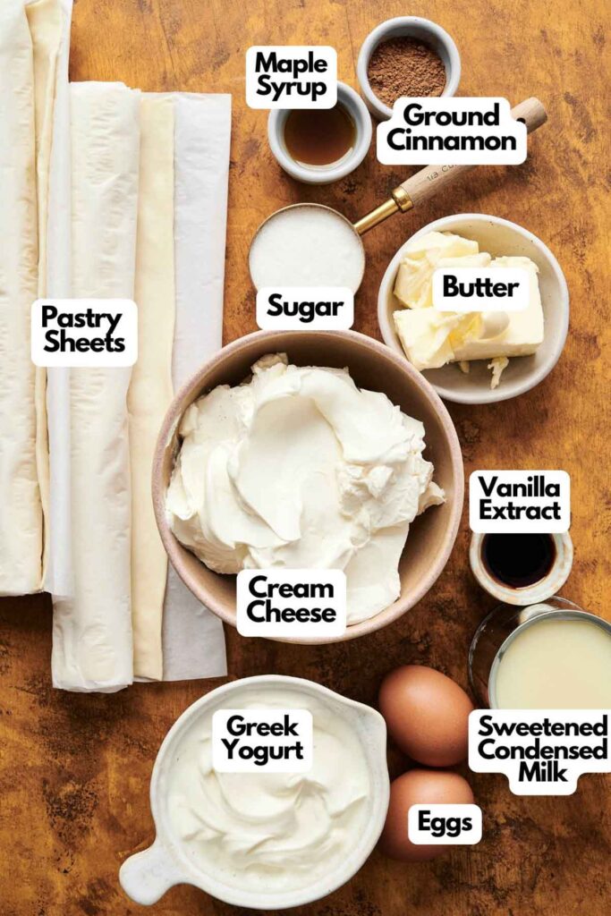 Assorted baking ingredients for a Churro Cheesecake, including pastry sheets, cream cheese, Greek yogurt, eggs, sweetened condensed milk, butter, sugar, maple syrup, ground cinnamon, and vanilla extract.