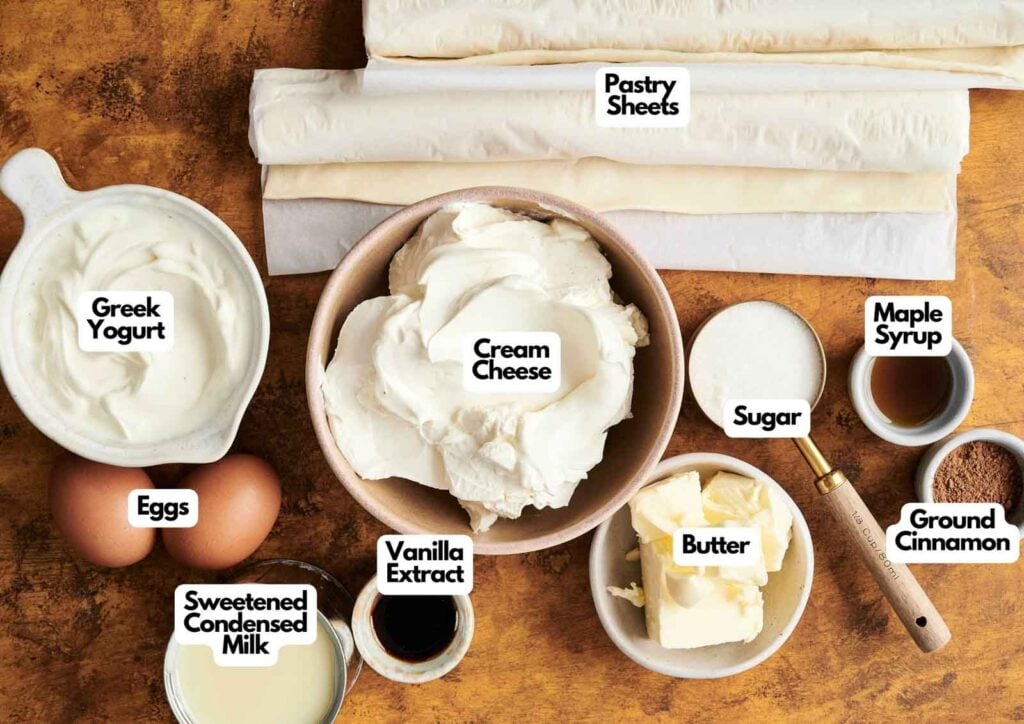 Various baking ingredients labeled, including cream cheese, pastry sheets, Greek yogurt, eggs, sweetened condensed milk, vanilla extract, butter, sugar, maple syrup, and ground cinnamon&mdash;perfect for making a delicious Churro Cheesecake.