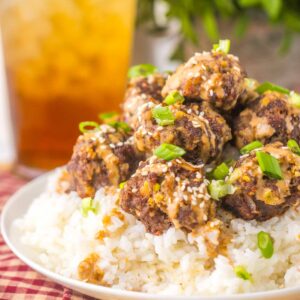 Plate of firecracker meatballs with sauce and green onions on white rice, accompanied by a tall glass of iced tea in the background.