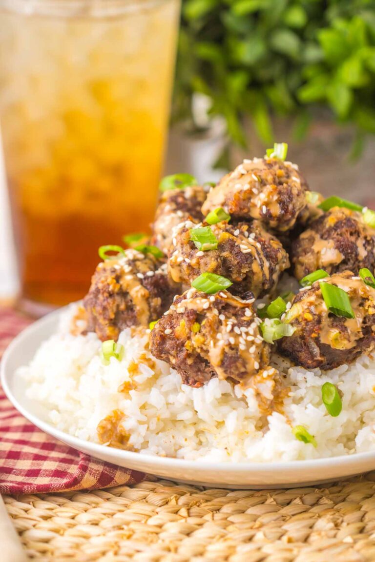 Plate of firecracker meatballs with sauce and green onions on white rice, accompanied by a tall glass of iced tea in the background.