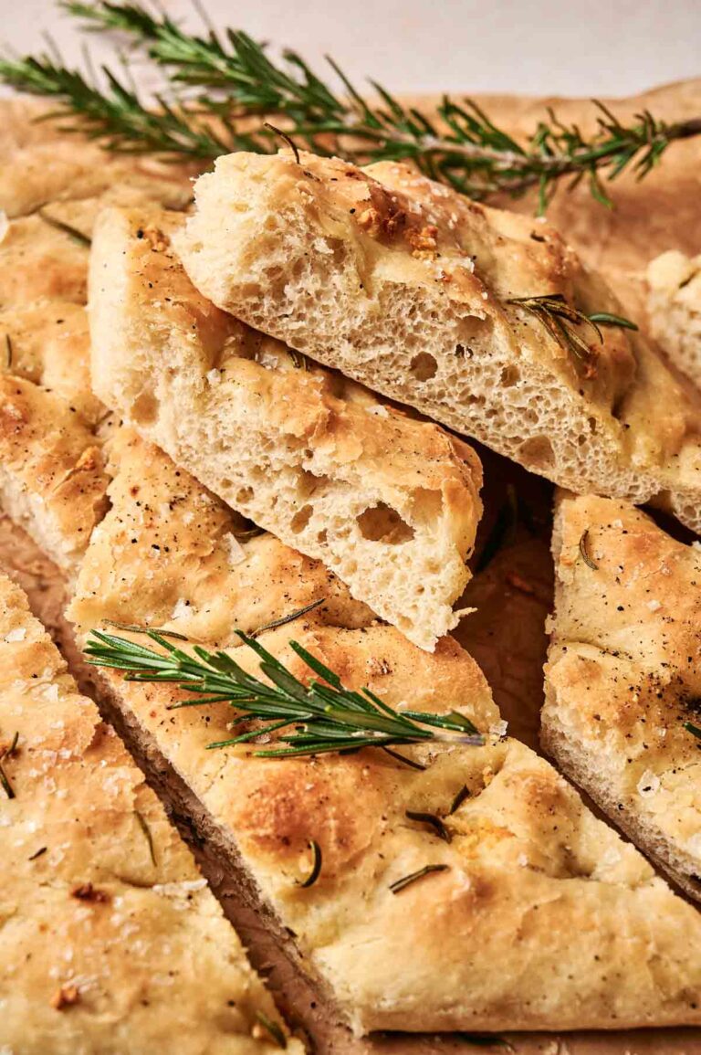 Slices of freshly baked focaccia bread topped with rosemary and herbs, arranged on brown paper.